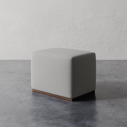 Lemieux et Cie Sophie Rectangular Ottoman in neutral upholstery with wood base, compact seating piece on concrete floor against textured plaster walls.