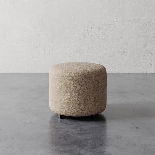 Lemieux et Cie Fabien Round Ottoman in neutral bouclé, cylindrical upholstered ottoman on concrete floor against textured plaster walls.