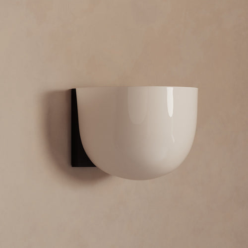 Lemieux et Cie Cheverny sconce with rounded white glass shade and dark wall mount, shown on neutral plaster wall in soft light.