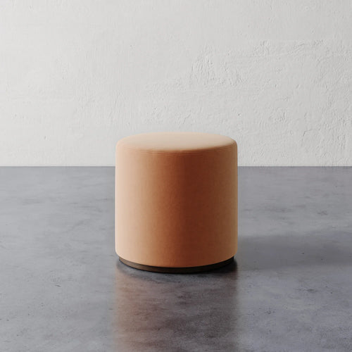 Lemieux et Cie Sophie Round Ottoman in warm peach upholstery, cylindrical ottoman on dark base, shown on concrete floor against textured plaster walls.