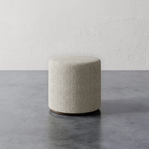 Lemieux et Cie Sophie Round Ottoman in ivory bouclé, cylindrical ottoman on dark wood base, shown on concrete floor against textured plaster walls.
