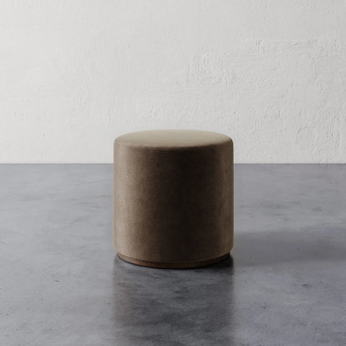 Lemieux et Cie Sophie Round Ottoman in warm taupe upholstery, cylindrical ottoman on polished concrete floor against textured plaster wall