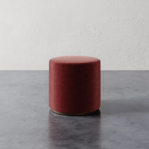 Lemieux et Cie Sophie Round Ottoman in terracotta upholstery, cylindrical ottoman on polished base, shown on concrete floor against textured plaster wall.