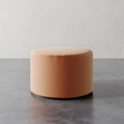 Lemieux et Cie Sophie Round Ottoman in warm blush upholstery, cylindrical ottoman on concrete floor against textured plaster wall.