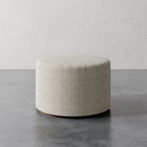 Lemieux et Cie Sophie Round 25" Ottoman in ivory bouclé, cylindrical ottoman on concrete floor against textured plaster wall.