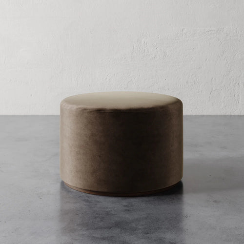 Lemieux et Cie Sophie Round Ottoman in warm taupe, low cylindrical upholstered ottoman on stone floor against textured plaster wall.