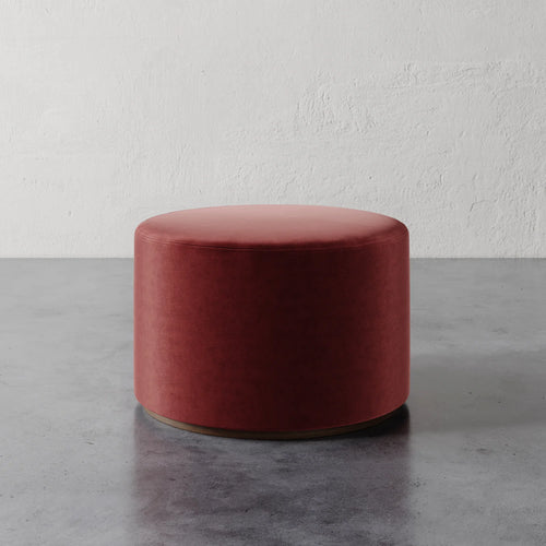 Lemieux et Cie Sophie Round Ottoman in terracotta velvet, cylindrical upholstered ottoman with rounded top, shown on polished concrete floor against textured plaster walls.
