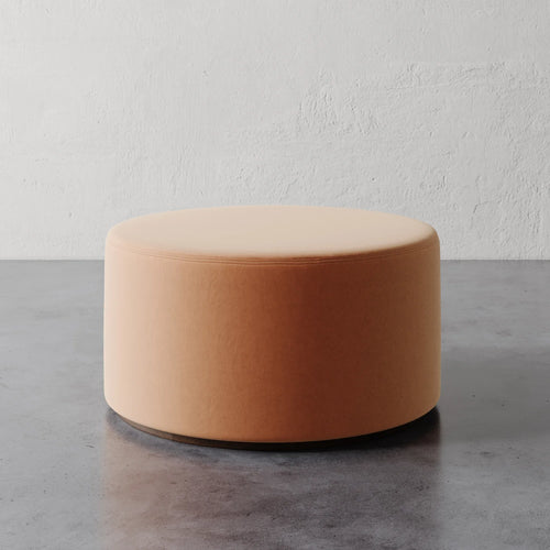 Lemieux et Cie Sophie Round Ottoman in blush upholstery, low-profile cylindrical ottoman on stone floor against textured plaster wall.