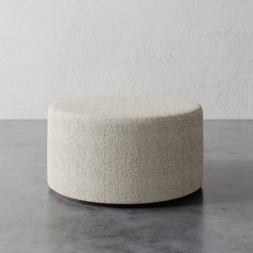 Lemieux et Cie Sophie Round 36" Ottoman in ivory bouclé, low-profile cylindrical ottoman on stone gray floor against textured plaster walls.