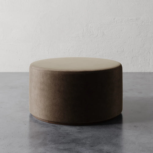 Lemieux et Cie Sophie Round 36" Ottoman in warm neutral upholstery, low cylindrical form on stone floor against textured plaster walls.