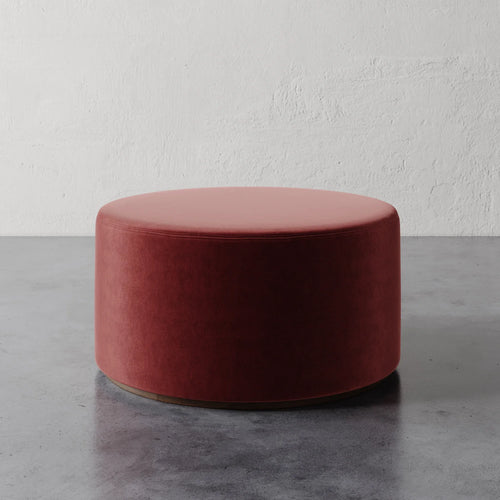 Lemieux et Cie Sophie Round 36" Ottoman in coral upholstery, low cylindrical ottoman on concrete floor against textured plaster wall.