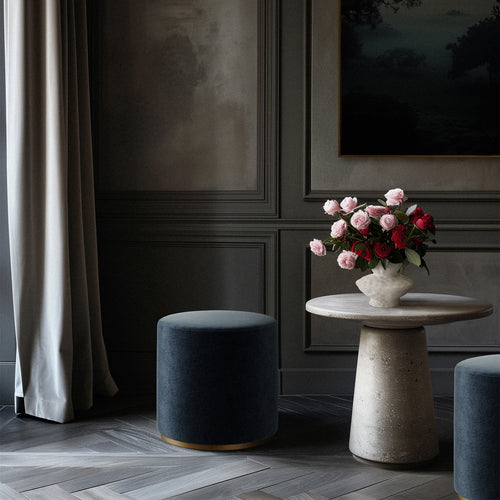 Lemieux et Cie Sophie Round Ottoman in charcoal upholstery with dark cylindrical ottomans and stone side table, styled in paneled sitting room with parquet floors.