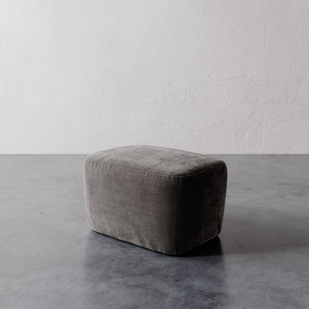 Lemieux et Cie Aurélie Ottoman in warm gray, low-profile rounded pouf shown on polished concrete floor against textured plaster walls.