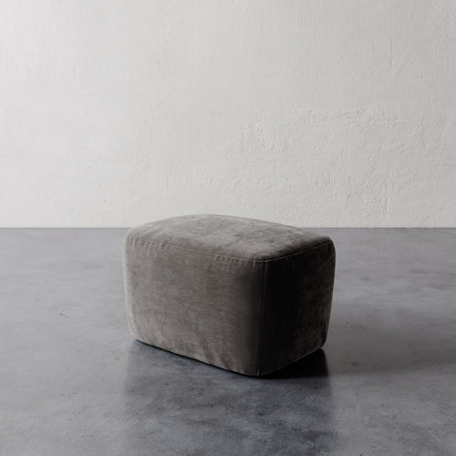 Lemieux et Cie Aurélie Ottoman in warm gray, low-profile rounded pouf shown on polished concrete floor against textured plaster walls.