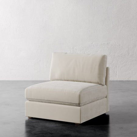 Lemieux et Cie Chanaux Armless Chair in cream upholstery, slipper chair with low-profile base, shown on dark concrete floor against textured white plaster wall.
