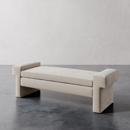 Lemieux et Cie Lemaire Bench in ivory upholstery, low-profile bench with sculptural block arms, shown on dark concrete floor against textured plaster walls.