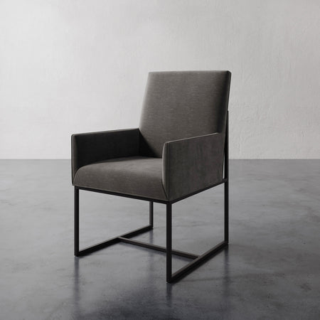 Lemieux et Cie Dupont Dining Arm Chair in charcoal upholstery with dark geometric base, shown on polished concrete floor against textured white wall.