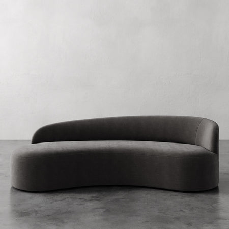 Lemieux et Cie Colette Right Arm Sofa in dark leather, curved asymmetric form with organic silhouette on concrete floor against plaster wall.