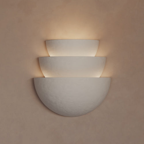 Lemieux et Cie Argan Sconce in ivory plaster, tiered half-moon wall light casting warm glow on neutral textured wall.