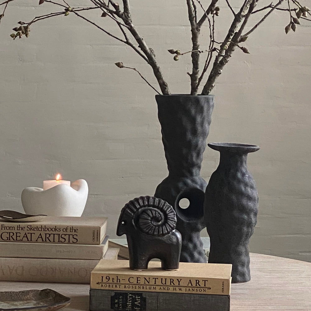 Lemieux et Cie Volcanic Vase in dark volcanic finish, sculptural organic vessel with branches, styled on stacked art books with candle and neutral wall.