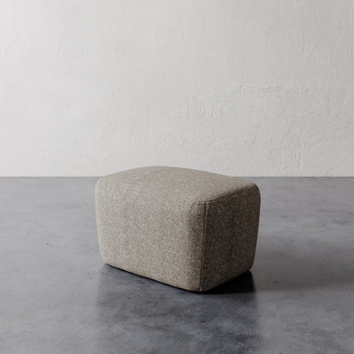 Lemieux et Cie Aurélie Ottoman, rounded low-profile pouf in warm grey, shown on concrete flooring against textured plaster walls.