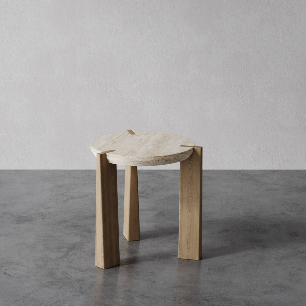 Lemieux et Cie Aline Side Table, round sculpted top with curved edges on four square legs, shown on polished concrete floor against soft gray wall.
