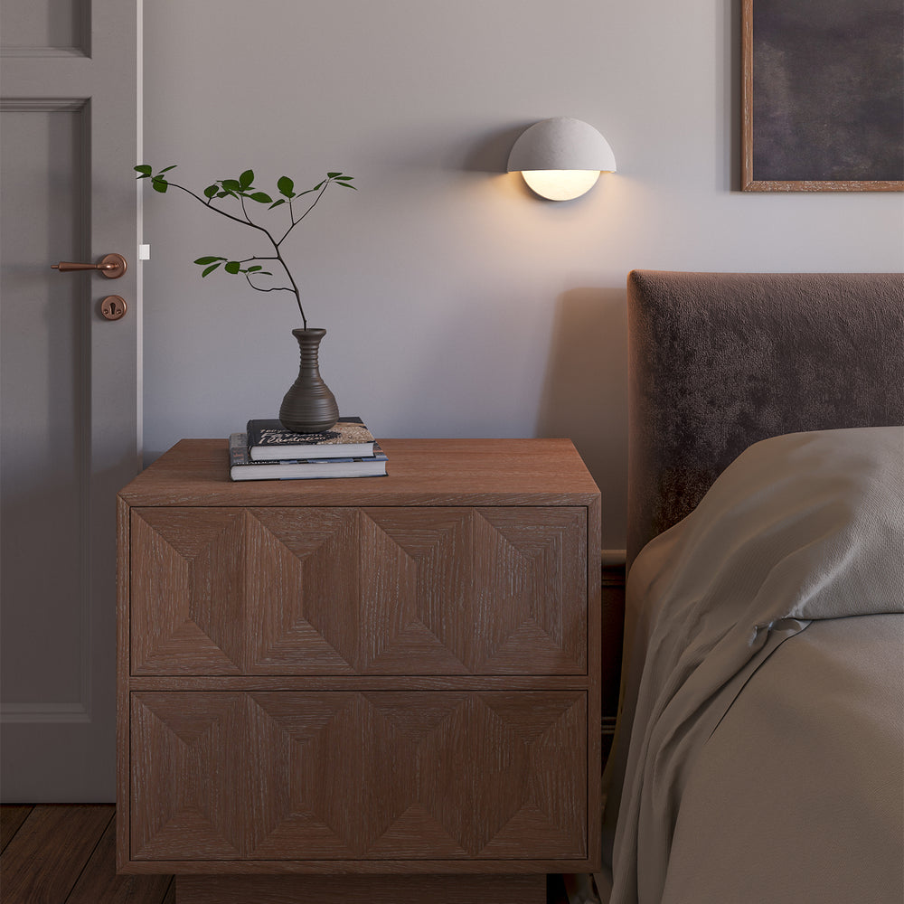 Lemieux et Cie Beaunay Small Sconce in steel and cast plaster, a designer lighting fixture above a bed corner with a nightstand, books, and vase in a sunlit European-style room.