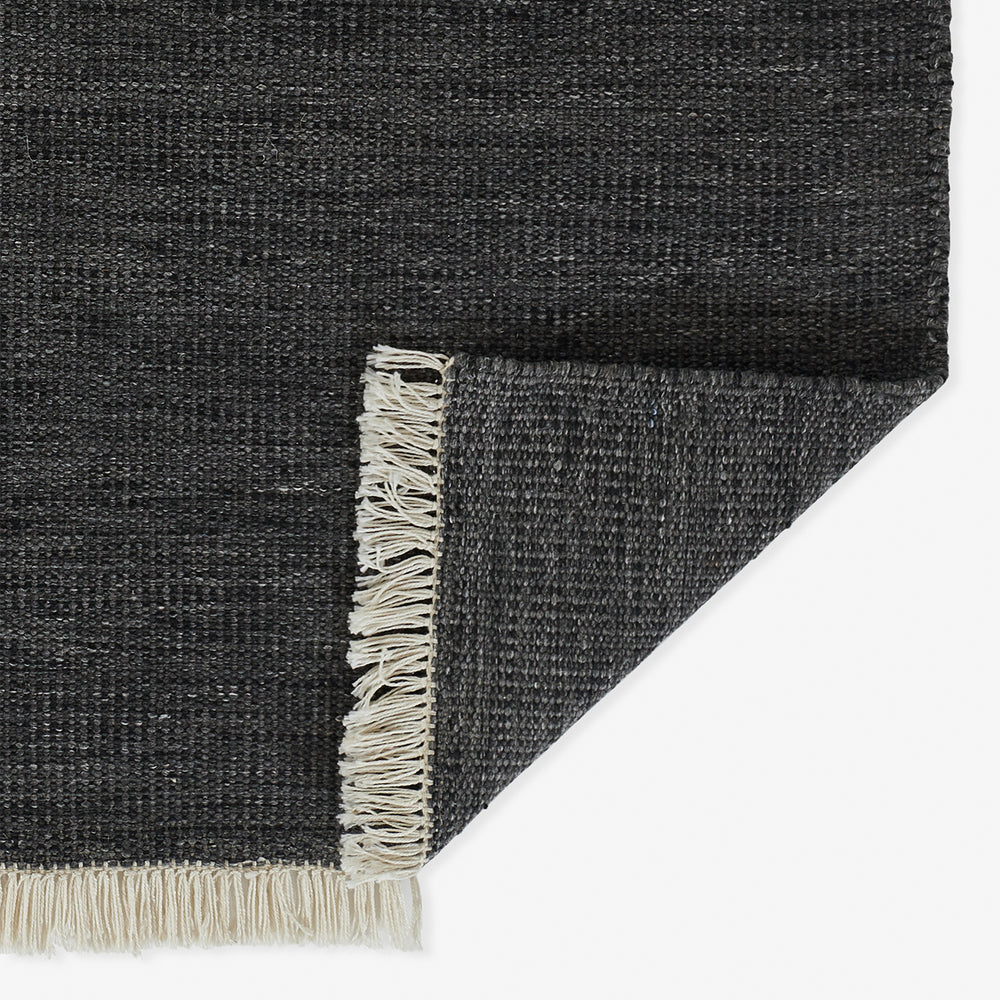 Lemieux et Cie Cove Chair in charcoal woven rug, handwoven flat-weave with natural fringe trim shown on white studio backdrop