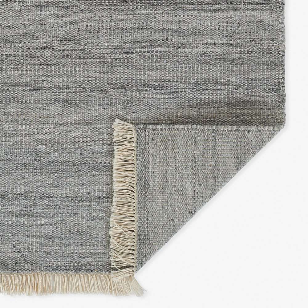 Lemieux et Cie handwoven rug in gray tones with natural fringe, corner detail showing textured surface and woven backing on white backdrop.
