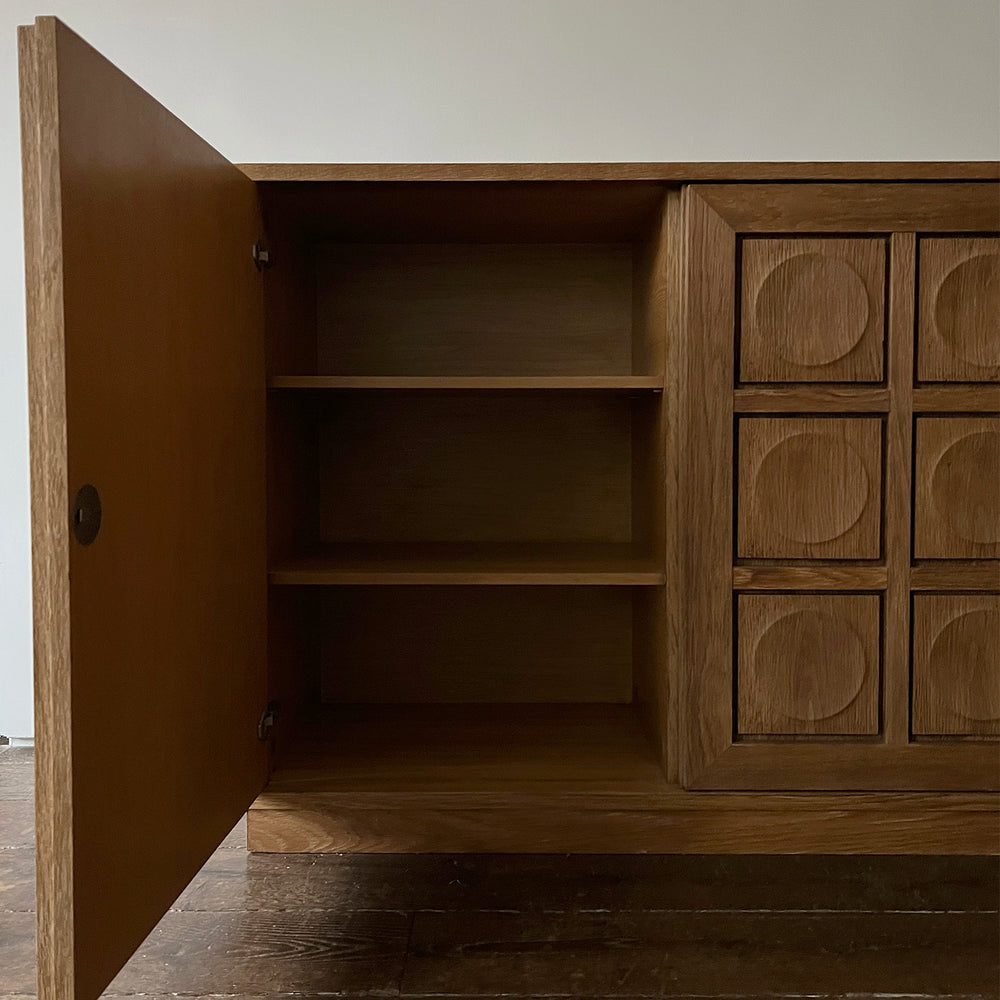 Lemieux et Cie platform sofa in cerused European oak with open cabinet door revealing interior shelving, geometric circle-paneled doors, dark wood flooring