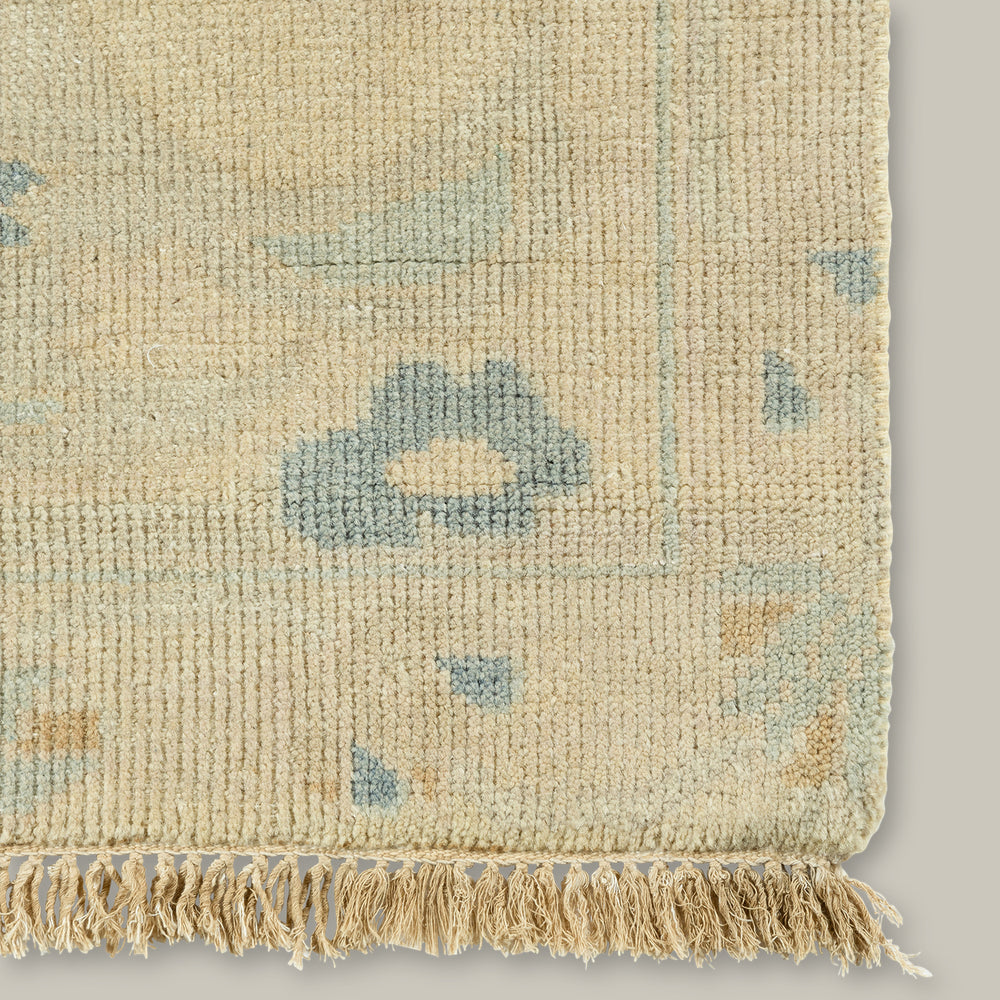 Lemieux et Cie Kouang rug corner detail in dusty neutral tones with soft blue accents, hand-knotted wool with fringe edge, shown on white studio backdrop.