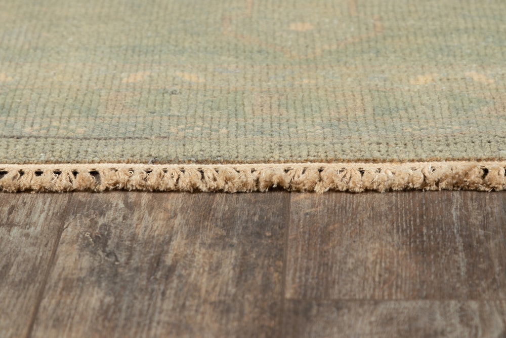 Lemieux et Cie Kouang rug edge detail in soft sage and warm tones, hand-knotted wool with whisper-thin pile, shown on weathered wood flooring.