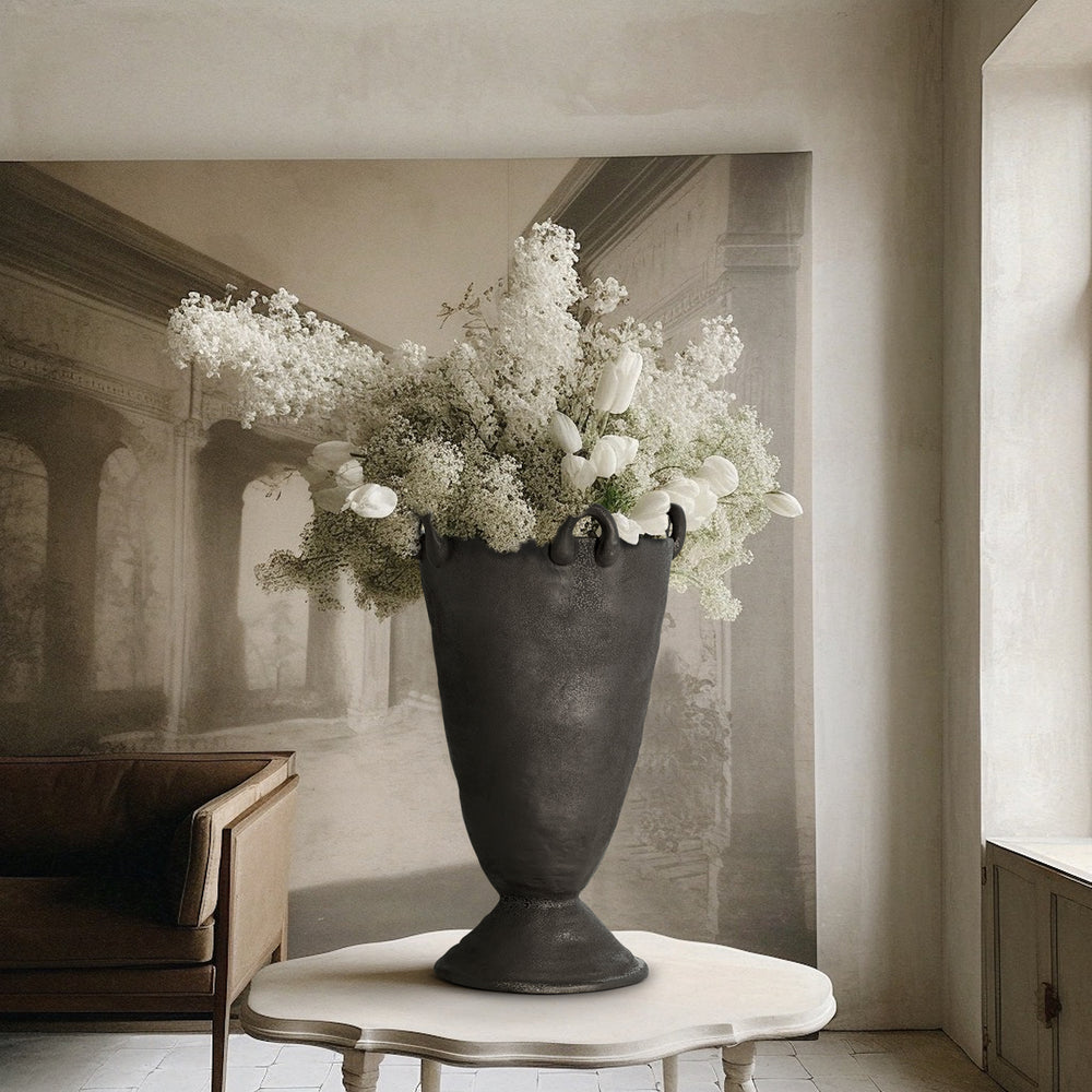 Lemieux et Cie Louis Vase in black, handcrafted footed urn with handles, atop a white marble table in a room with neutral plaster walls.