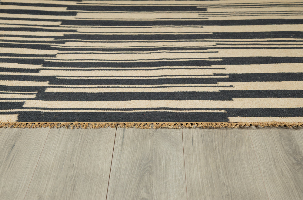 Lemieux et Cie Duar rug in charcoal and ivory striped pattern, handwoven flatweave with fringe edge, on pale wood flooring.