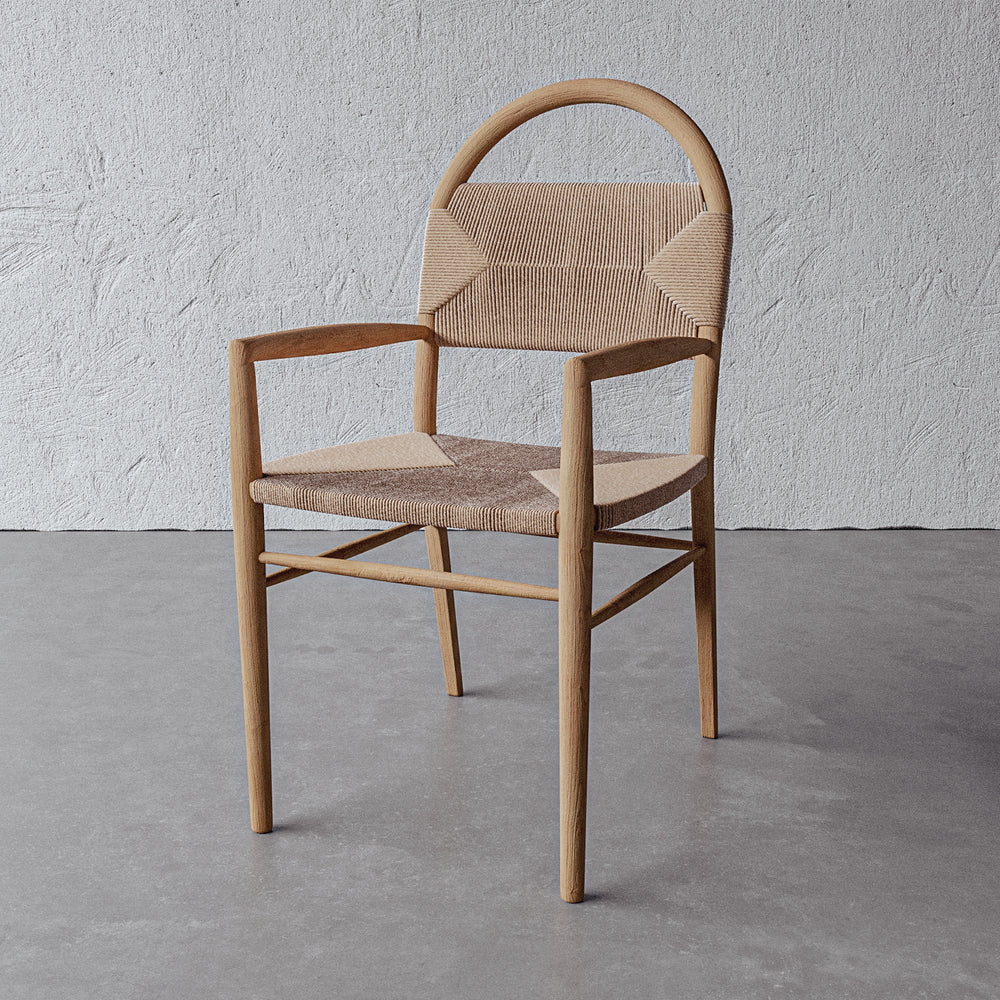 Lemieux et Cie Pernelle Dining Arm Chair in oak with woven rush seat and arched backrest, shown on concrete floor against white textured plaster wall.
