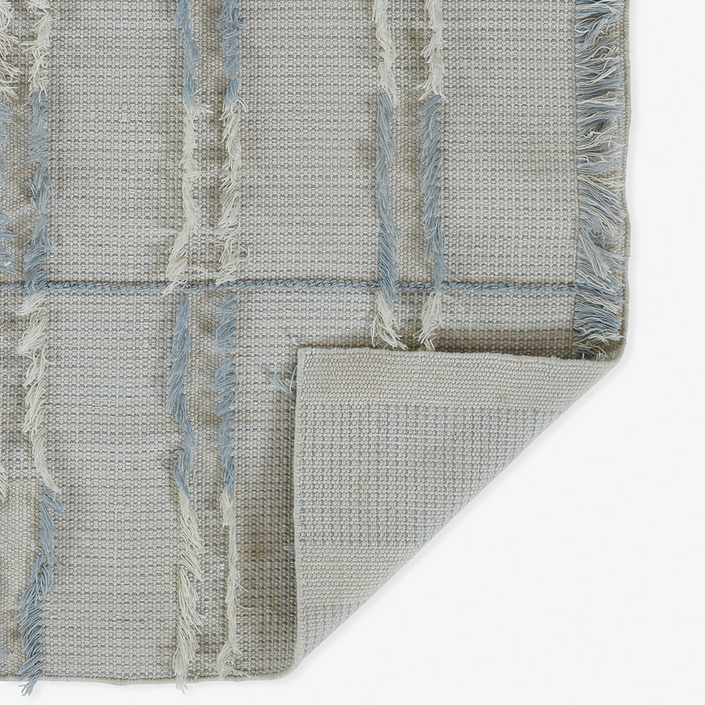Lemieux et Cie Cluny rug in neutral tones with blue windowpane grid pattern, hand-woven wool with fringe detailing, shown on white studio backdrop with corner folded to reveal reverse