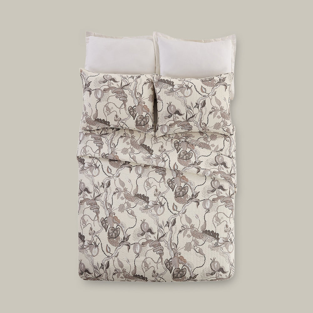 Lemieux et Cie Snake Floral Quilt in white, taupe and gray on cotton, channel-stitched bedding with serpent motif, styled on a bed with white pillows against neutral wall.