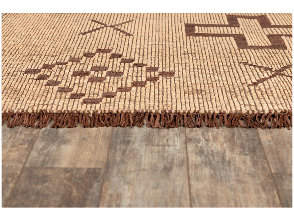 Lemieux et Cie Tugart Rug in tan and brown handwoven leather and jute with geometric pattern and leather fringe, shown on weathered wood flooring.
