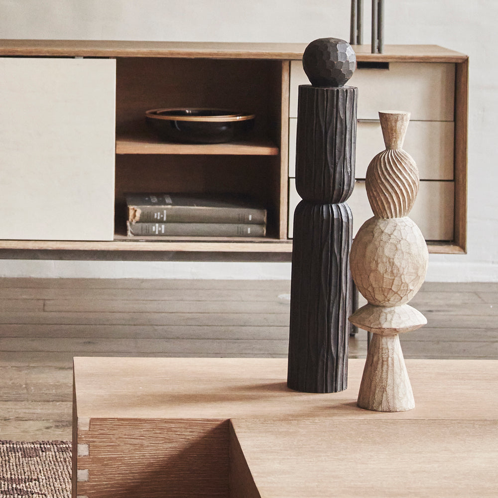 Lemieux et Cie Amelot Pillar, sculptural dark wooden column with fluted details and faceted sphere, displayed on light wood flooring near a low cabinet in neutral interior.