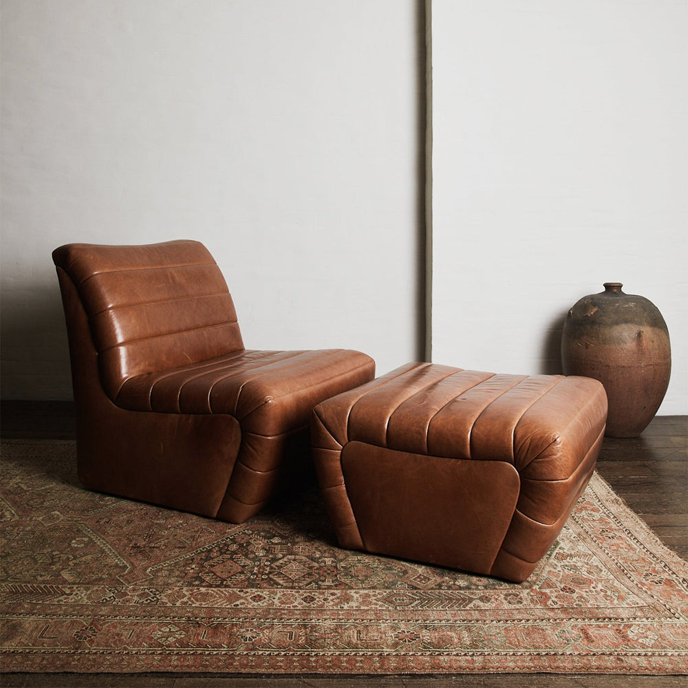 Lemieux et Cie channeled leather lounge chair and ottoman with ribbed upholstery, shown on vintage rug atop dark wood floors with ceramic vase and white walls.