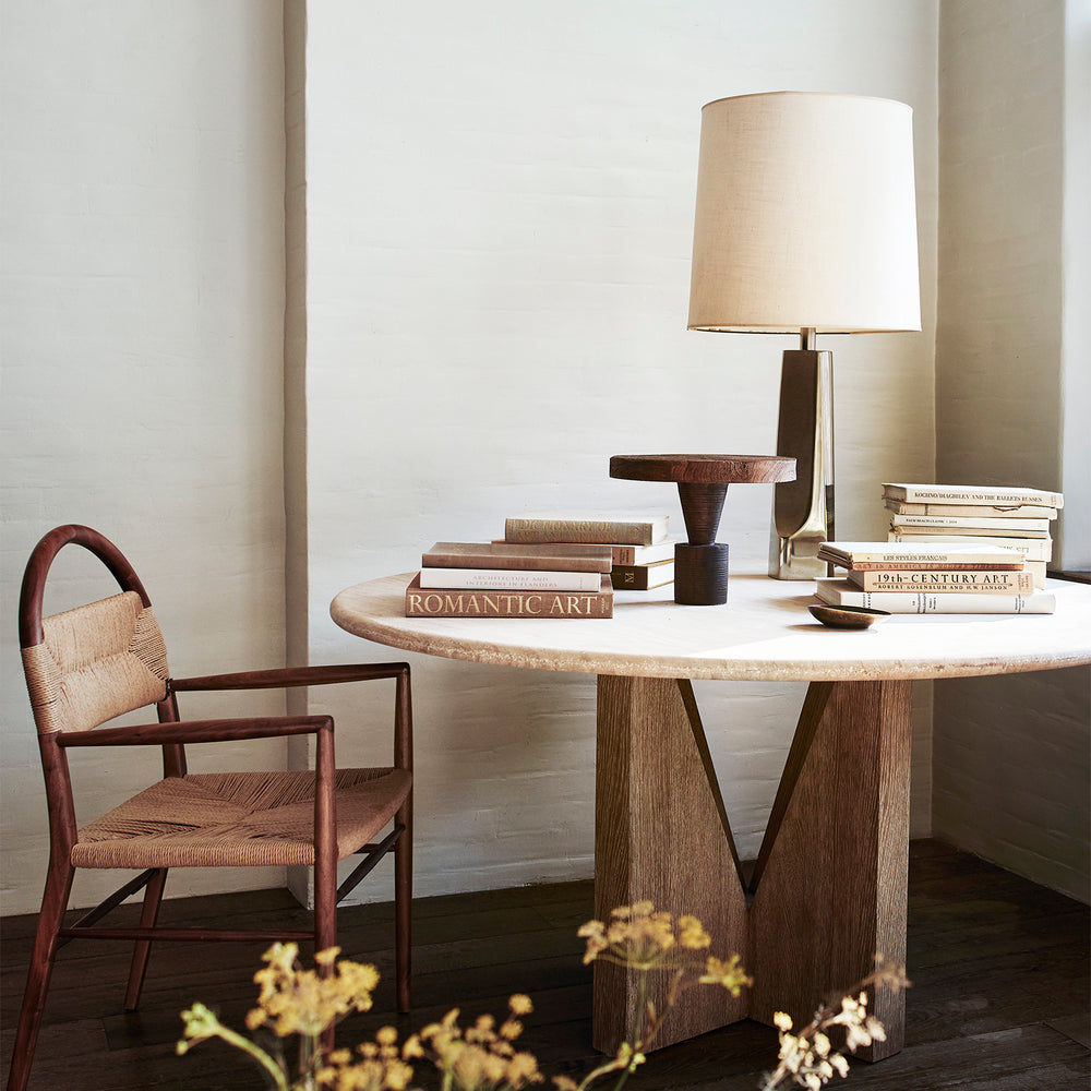 Lemieux et Cie Damiette Pedestal, round table with stone top and geometric wood base, styled with art books and table lamp in neutral sitting room with dark wood floors.
